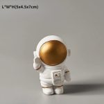 Miniatura de astronauta dourado com capacete grande, detalhes em branco e uma mochila de suporte, em pose de saudação.