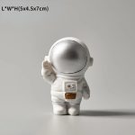 Miniatura de astronauta com capacete dourado, traje branco e detalhes em dourado, em posição de cumprimento, com fundo neutro.