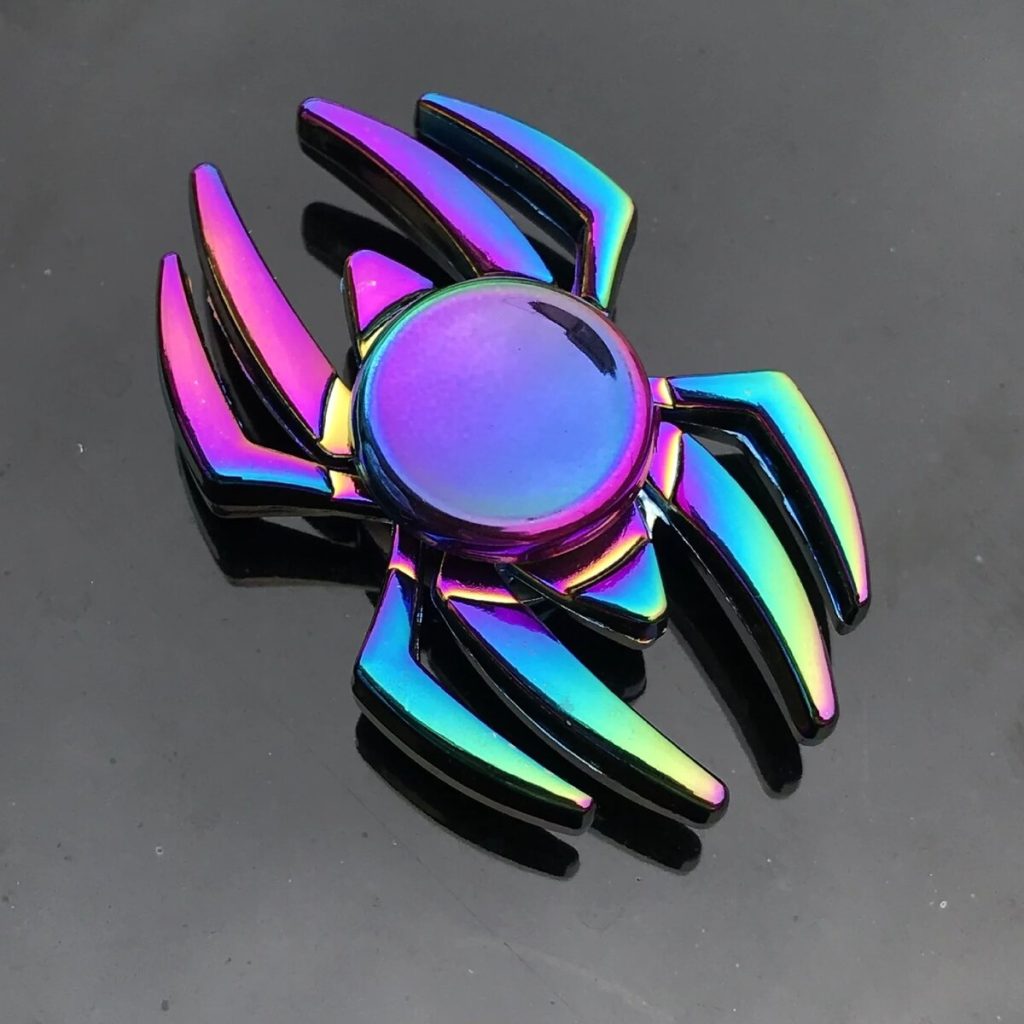 Fidget spinner com corpo de aranha em cores iridescentes, com oito patas e uma superfície lisa no centro, colocado sobre uma superfície escura.