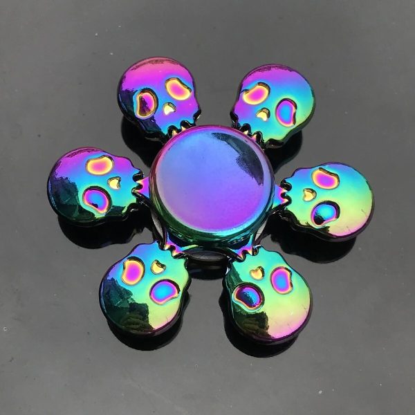 Fidget spinner com design de caveira, acabamento holográfico com cores iridescentes, visto de cima sobre fundo escuro, detalhes brilhantes e reflexivos.