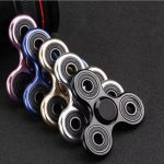 Conjunto de cinco fidget spinners em cores metálicas, com diferentes acabamentos, colocados sobre uma superfície escura.