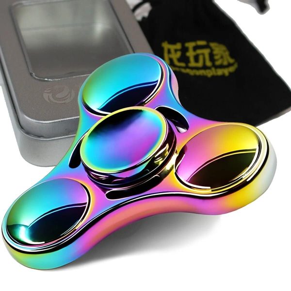 Fidget spinner de metal com acabamento iridescente, mostrando detalhes das três ranhuras e o centro rotativo, com embalagem de transporte ao fundo.