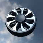 Fidget spinner com design de roda de carro, com oito pás pretas e brancas, visto de cima sobre uma superfície refletora.