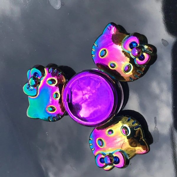 Fidget spinner com forma de gato, acabamento holográfico que reflete várias cores, com detalhes de olhos e orelhas, colocado sobre uma superfície escura.
