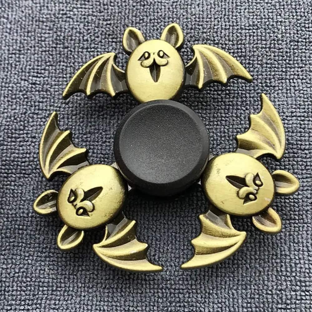 Fidget spinner com detalhes de morcego, asas e rosto sorridente, em cores douradas e pretas, colocado sobre tecido cinzento.