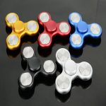 Fidget spinners de metal em cores dourado, vermelho, azul, preto e branco, dispostos sobre uma superfície preta, com reflexos visíveis.