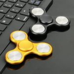Dois fidget spinners, um dourado e um preto, colocados ao lado de um teclado de computador, destacando o design circular e detalhes metálicos.