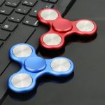 Dois fidget spinners, um vermelho e um azul, colocados sobre um teclado de computador, destacando os detalhes metálicos e o design ergonómico.