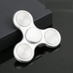 Fidget spinner branco com três braços e centro giratório, colocado sobre uma superfície escura com caixa preta ao fundo.