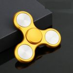 Fidget spinner dourado com três braços e centro circular, colocado sobre uma superfície escura ao lado de uma caixa preta.