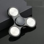 Fidget spinner preto com três braços e acabamento metálico, colocado sobre uma superfície escura com caixa ao fundo.