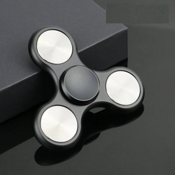 Fidget spinner preto com três braços e acabamento metálico, colocado sobre uma superfície escura com caixa ao fundo.