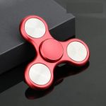 Fidget spinner de cor vermelha com três braços, cada um com uma roda de metal, colocado sobre uma caixa preta.