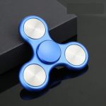 Fidget spinner azul com três braços e rolamentos metálicos, colocado sobre uma superfície preta com caixa escura ao fundo.