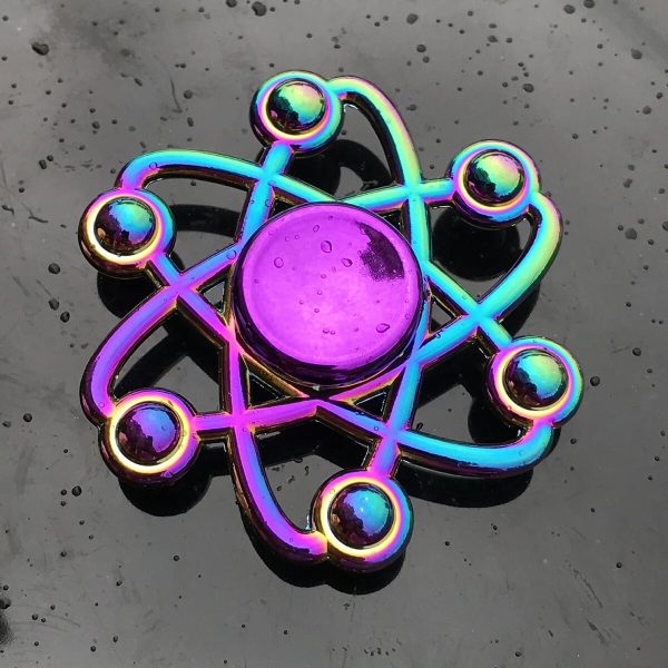 Fidget spinner com forma de átomo, com núcleo central roxo e braços metálicos brilhantes com esferas nas extremidades, sobre fundo escuro.
