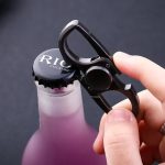Ferramenta de metal preta com alavanca, usada para abrir garrafas, sendo segurada por uma mão com unhas curtas e limpas, contra fundo escuro.