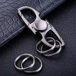 Chaveiro com design de fidget spinner em metal, com anéis de chave adicionais ao lado, colocado sobre uma superfície preta com textura semelhante a couro.