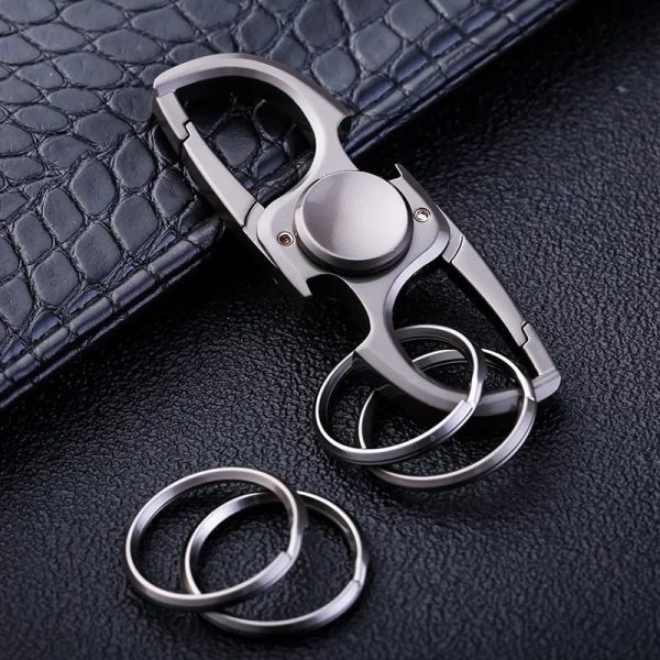 Chaveiro com design de fidget spinner em metal, com anéis de chave adicionais ao lado, colocado sobre uma superfície preta com textura semelhante a couro.