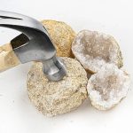 Geodes de ágata cortadas ao meio, mostrando cristais brancos e cinzentos no interior, com martelo de escultura ao lado.
