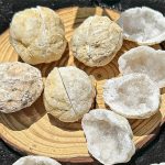 Seis geodes de ágata fechados e abertos, mostrando cristais brancos e interior cristalino, dispostos numa bandeja de madeira.