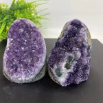 Duas metades de geodo de ametista, uma com cristais roxos brilhantes e outra com cristais escuros e inclusões, sobre fundo neutro com plantas verdes ao fundo.