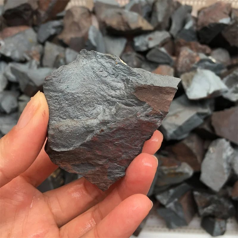 Pedra de hematite bruta com superfície irregular, cinza escuro e brilho metálico, segurada por uma mão, com várias pedras semelhantes ao fundo.