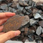 Pedra de hematite em bruto, com superfície metálica e textura irregular, sendo segurada por uma mão, com fundo de várias outras pedras semelhantes.