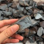 Pedaço de hematite bruto com superfície escura e textura irregular, sendo segurado por uma mão, com uma pilha de minerais semelhantes ao fundo.