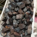 Vários pedaços de hematite em bruto, com superfícies irregulares e cores cinza escuro a preto, dispostos numa caixa de plástico branca.