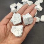 Três pedaços de Howlite branca com veios cinzentos, segurados na mão, com mais pedaços ao fundo.