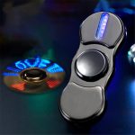 Fidget spinner com luz LED azul acesa, colocado ao lado de um disco de luz colorido em fundo escuro.