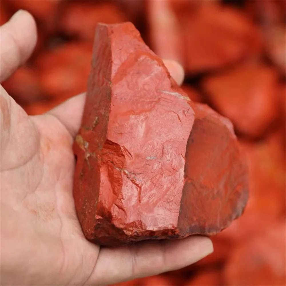 Pedra de jaspe vermelho com superfície irregular e cores vibrantes, vista de perto na mão de uma pessoa, com fundo de outras pedras semelhantes.