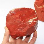 Pedra de jaspe vermelho em bruto, com textura irregular e cores vibrantes, sendo segurada por uma mão, com outra pedra ao fundo.