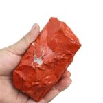 Pedra de jaspe vermelho com superfície irregular, mostrando textura natural e cores vibrantes, sendo segurada por uma mão humana.