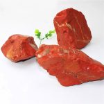 Três rochas de jaspe vermelho com veios dourados, uma delas com uma pequena planta verde ao lado, sobre fundo branco.