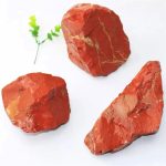 Três pedaços de jaspe vermelho com veios dourados, diferentes formas e tamanhos, sobre fundo branco, com uma pequena planta decorativa ao lado.
