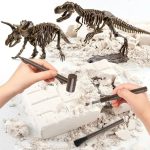 Duas mãos a usar ferramentas de escavação em uma caixa de areia com fósseis de dinossauros e esqueletos de dinossauros ao fundo.