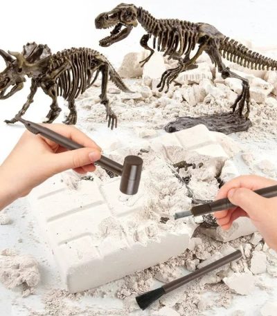 Duas mãos a usar ferramentas de escavação em uma caixa de areia com fósseis de dinossauros e esqueletos de dinossauros ao fundo.