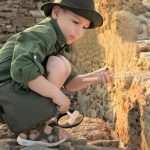 Menino com chapéu e roupa de explorador a examinar pedras antigas com uma ferramenta de escavação.