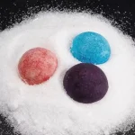 Três bolas de sal coloridas (vermelha, azul e roxa) dispostas sobre uma camada de sal branco, com fundo preto.