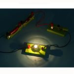 Circuito elétrico com lâmpada acesa, fios vermelhos e verdes, componentes de plástico amarelo e base de suporte.