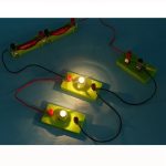Circuito elétrico com duas lâmpadas acesas, fios vermelhos e pretos, e componentes de plástico verde, demonstrando funcionamento de circuito simples.