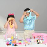 Duas crianças brincando com slime de gelado, uma menina com laço rosa e um rapaz com camisola azul, rodeados de ingredientes e utensílios de slime.