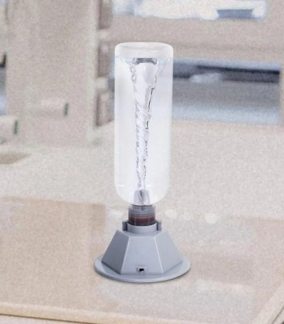 Tubo de vidro com tornado visível dentro, apoiado numa base cinzenta, em ambiente de laboratório ou sala de aula.