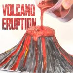 Reprodução de um vulcão com lava de cor vermelha a escorrer, com embalagem do kit visível ao lado, usado para demonstrações educativas de erupções vulcânicas.
