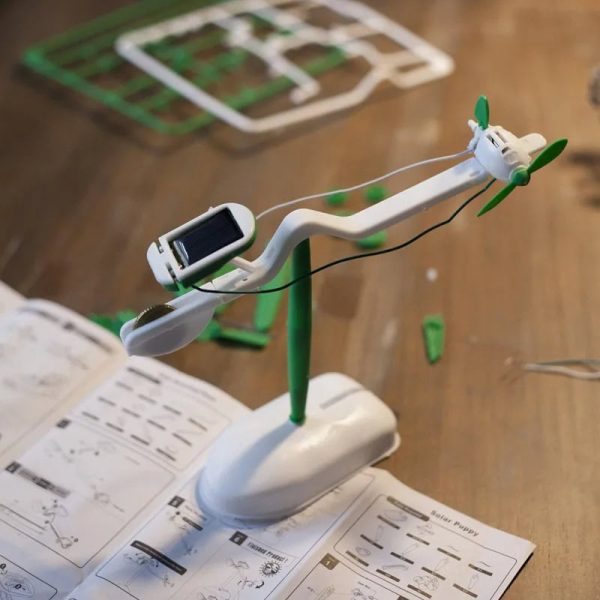 Pequeno veículo movido a energia solar com hélices verdes, montado em suporte branco, com painéis solares e componentes visíveis, sobre mesa de trabalho.