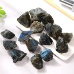 Pedras de labradorite bruta com reflexos azuis e verdes, variadas formas e tamanhos, sobre uma superfície branca.
