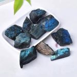 Pedras de labradorite bruta com cores azuladas e verdes, variadas formas e tamanhos, sobre prato branco com fundo claro.
