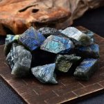 Pedras de labradorite bruta exibindo cores azuis e verdes com efeito iridescente, sobre uma base de madeira escura, com fundo de madeira natural.