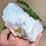 Pedra de larimar em bruto, com tonalidades azuis e detalhes de textura, vista de perto na mão, com folhas verdes ao fundo.